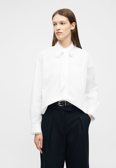 Femme portant une chemise blanche à manches longues avec un col pointu et un pantalon noir taille haute, debout devant un fond clair uni.