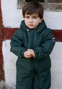 Tuta da neve impermeabile verde scuro con zip frontale, polsini elastici e due bottoni decorativi in bronzo. Bambino in piedi contro una parete testurizzata.