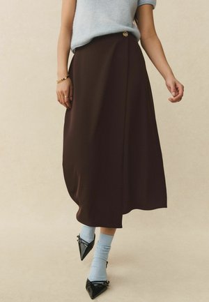 Frau trägt ein hellblaues ärmelloses Oberteil, einen dunkelbraunen Midirock mit Knopfdetails, hellblaue Socken und schwarze spitze Schuhe.