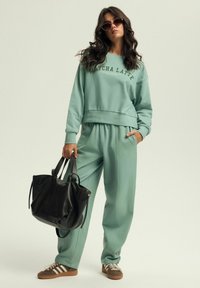 Miętowy zestaw dresowy z bluzą i spodniami joggers, z prążkowanymi mankietami, z napisem "Matcha Latte". Czarna skórzana torba na ramię i brązowe sneakersy w paski.