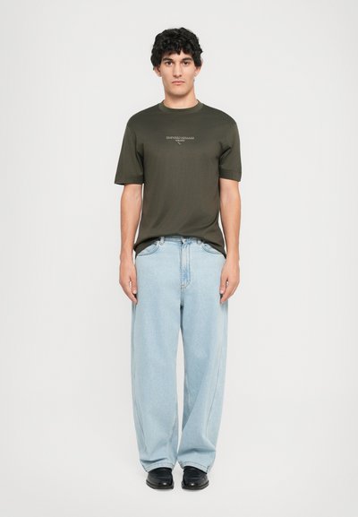 Fiatal férfi áll, olivazöld Emporio Armani pólót, világoskék, bő szárú farmert és fekete cipőt visel egy sima háttér előtt.