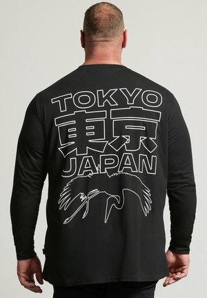 Hombre vistiendo una camiseta negra de manga larga con texto blanco "TOKYO" y "JAPAN" y caracteres japoneses encima de un contorno blanco de un pájaro grulla en la espalda.