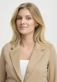 Blazer beige con un design strutturato, dotato di un colletto a rotondo e una texture liscia, abbinato a un top bianco a coste sottostante.