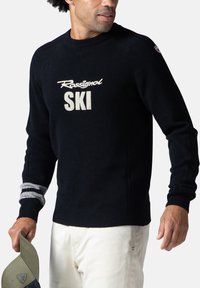 Maglione nero a maglia con logo "Rossignol SKI" in crema. Presenta polsini a coste con strisce grigie e scollatura rotonda. Indossato con pantaloni chiari.