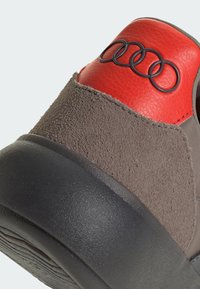 Teniska s hnědým semišovým a červeným koženým svrškem, černou gumovou podrážkou a výrazným logem Audi na patě.