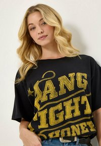 Musta ylipitkä t-paita, jossa on rohkeaa keltaista tekstiä ja narukuvioita. Suunniteltu lyhyillä hihoilla ja rentoa istuvuutta varten.