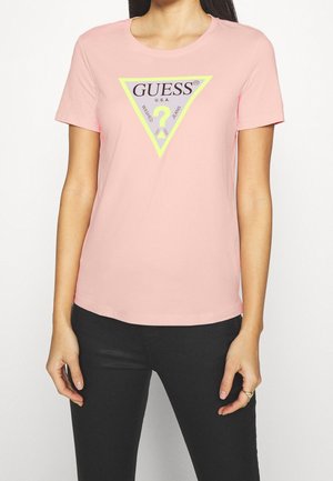 Kobieta w różowej koszulce z krótkim rękawem, na której znajduje się trójkąt z żółtą obwódką z napisem „GUESS U.S.A.” i logo z pytajnikiem na środku.