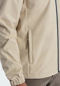 Beige, leichte Jacke mit strukturiertem Design, Reißverschluss, elastischen Bündchen und einer Seitentasche. Glatte Textur und minimales Detail.