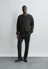 Dark green fleece sweatshirt with a round neckline, paired with black tailored trousers and black shoes, set against a plain light background.