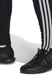 Zwarte sportieve schoenen met een geglazuurd patroon en zwarte veters, gecombineerd met zwarte broek met aan de zijkanten drie witte strepen.
