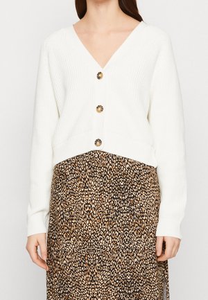 Frau mit weißem, geripptem Cardigan mit Schildpattknöpfen über einem braunen und schwarzen Leopardenmusterrock.