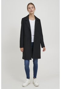 Cappotto nero con colletto a rever, due bottoni e tasche laterali. Indossato sopra una camicia bianca e jeans blu, abbinato a sneakers bianche.