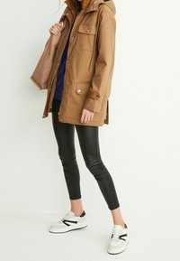 Brun parka jakke med hætte, med forlommer og metaltrykknapper, parret med sorte leggings og hvide sneakers med sorte detaljer.