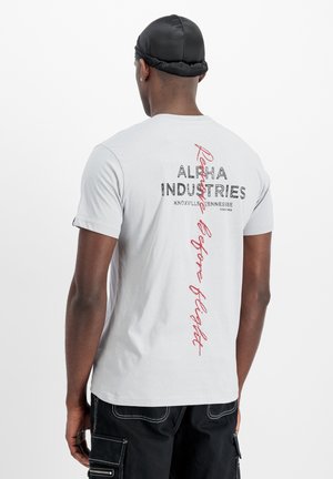 Mand iført hvid Alpha Industries T-shirt med sort og rød tekst på ryggen, sorte shorts og sort kasket, set bagfra mod hvid baggrund.