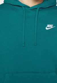 Tyrkysová mikina Nike s kapsou na kangura vpředu, s tkaničkami a bílým logem vyšitým na levé hrudi. Měkká textura, neformální design.