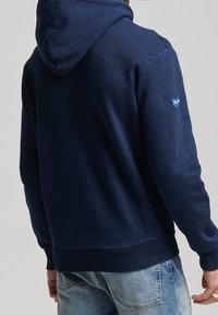 Hoodie blu navy con una texture morbida, presenta un cappuccio con cordino, polsini e orlo ribattuti, con una patch del logo sulla spalla sinistra.