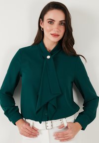 Dark green blouse with a collar and a large front tie detail. Features a button closure and long sleeves, paired with a white belt.