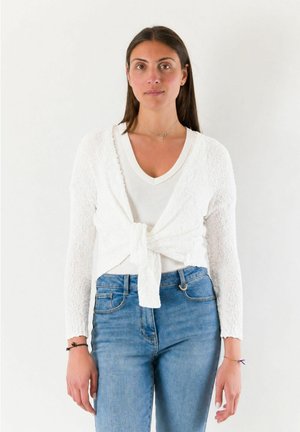 Donna con lunghi capelli scuri indossa un top bianco a scollo a V, un cardigan bianco strutturato legato in vita e jeans blu, in piedi davanti a uno sfondo semplice.