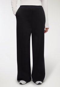 Pantaloni neri a gamba larga con una texture liscia e una cintura elastica, dotati di tasche laterali e abbinati a sneakers grigie.