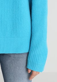 Pull en tricot turquoise avec une texture côtelée, présentant des manches longues et un ourlet large. Associé à un jean en denim clair.