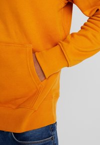 Persona vestida con sudadera naranja y una mano dentro del bolsillo delantero, combinada con jeans de denim azul, contra un fondo simple.