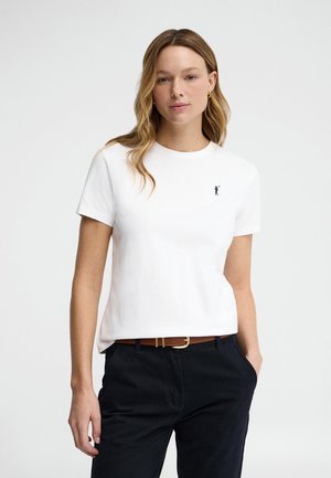 Vrouw met lang golvend haar die een wit T-shirt met korte mouwen en een klein donker logo draagt, zwarte broek en een bruine riem, staand met één hand in de zak.