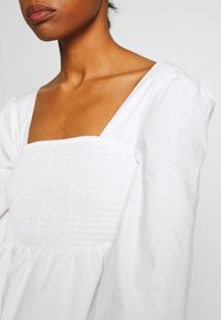 Vit blus med fyrkantig halsringning, smockat liv och puffärmar. Tillverkad av ett lätt tyg med en slät textur.