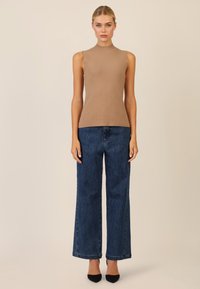 Ärmelloses, sandfarbenes Oberteil mit hohem Kragen; kombiniert mit dunkelblauen weit geschnittenen Jeans mit dezenten Verzierungen; schwarze spitze Absätze runden das Outfit ab.