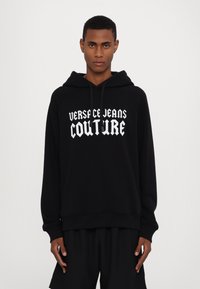 Μαύρη φούτερ μπλούζα με τσέπη καγκουρό, που φέρει το κείμενο "VERSACE JEANS COUTURE" σε λευκή γκόθικ γραμματοσειρά. Μείγμα βαμβακιού, άνετη εφαρμογή και κουκούλα με κορδόνι.