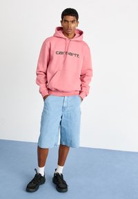 Sweat-shirt rose avec capuche et poche avant, arborant le logo "carhartt." en noir, associé à un short en denim bleu clair et des sneakers noires.