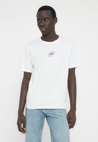 Valkoinen puuvillainen t-paita, jossa on liila ja sininen logoplässi rinnassa, lyhyet hihat ja klassinen pyöreä kaulus. Yhdistetty vaaleansinisiin farkkuihin.