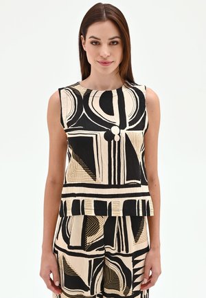 Femme portant un haut sans manches et un pantalon avec des motifs abstraits géométriques noirs et beiges sur fond blanc uni.