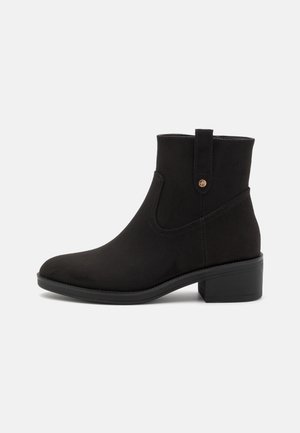 Sort ruskind ankle-støvle med lav blokhæl, rundet tå og en trækflig med en lille metallisk knap på siden.