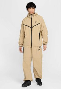 Coupe-vent beige Nike avec des accents noirs, doté d'une capuche, d'un zip avant et de poches latérales, associé à un pantalon ample assorti et à des chaussures noires.