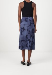 Žena stojí zády, oblečená v černém bezrukávovém topu, modré květinové midi sukni a černých klouzavých botách na bílém pozadí.