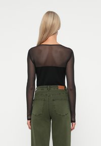 Čierny top s dlhým rukávom z priesvitného materiálu so solidnou zadnou časťou, skombinovaný s olivovozelenými nohavicami s vysokým pásom, ktoré majú vrecká a značkovú etiketu.