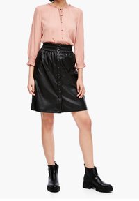 Blouse rose à manches longues avec un col noué et une patte de boutonnage sur le devant, associée à une jupe en fausse cuir noire avec une taille à volants et des détails à boutons.