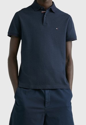 Homme portant un polo bleu marine avec un petit logo sur la poitrine et un short bleu marine assorti, les mains dans les poches.