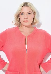 Chaqueta coral con cremallera en tejido suave, con mangas largas, cuello redondo y dos bolsillos frontales, con una textura lisa y cremallera plateada.