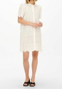 Vestito bianco a maniche corte con chiusura a bottoni, caratterizzato da motivi ricamati a occhio di pernice e orlo scallop. Indossato con sandali neri.