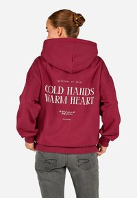 Bordo spalvos gobtuvė su dideliu baltu užrašu "ŠALTOS RANKOS, ŠILTA ŠIRDIS" ir mažesniais tekstais. Medvilnės mišinys, erdvus gobtuvas ir raštuoti rankogaliai.