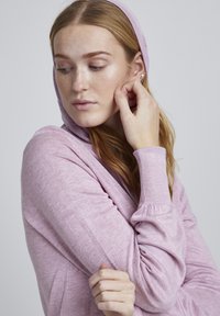 Haut à capuche rose clair avec une texture douce, des manches longues, des poignets côtelés et une capuche drapée. Comprend des détails de couture sur les manches.