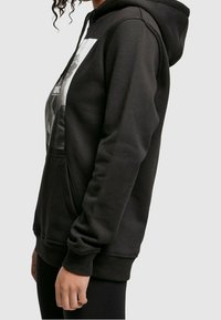 Osoba v černé mikině s kapucí a přední kapsou s grafickým potiskem, zobrazená z boku na jednoduchém pozadí.