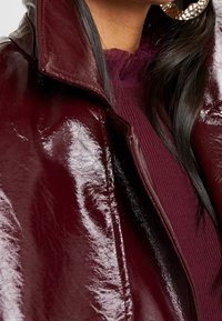 Burgunderfarbene Lacklederjacke mit hohem Kragen, die eine glänzende Textur aufweist. Darunter ist ein dunkelroter, gerippter Pullover teilweise sichtbar.