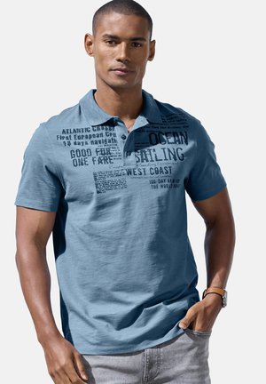 Young man wearing a blue polo shirt with dark nautical-themed text, standing with one hand in pocket against plain light background.