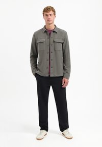 Grijze button-up overhemd met twee borstzakken, gedragen over een bordeauxrood shirt, gecombineerd met zwarte broek en witte sneakers. Effen achtergrond.