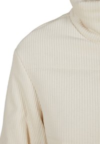 Pull à col roulé en tissu côtelé crème, montrant l'épaule et le haut de la manche avec des lignes texturées verticales.
