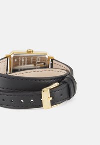Montre avec un bracelet en cuir noir et un boîtier rectangulaire en ton or. Elle présente une surface polie, une boucle ajustable et des trous de clasp visibles sur le bracelet.