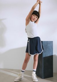 Marineblauwe sportshorts met witte bies, elastische tailleband en zijzakken, gecombineerd met een witte mouwloze top en witte hoge sneakers.