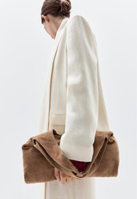 Clutch bag in suede marrone con design pieghettato, tenuto con una mano, abbinato a un blazer beige chiaro e a un maglione rosso sotto.